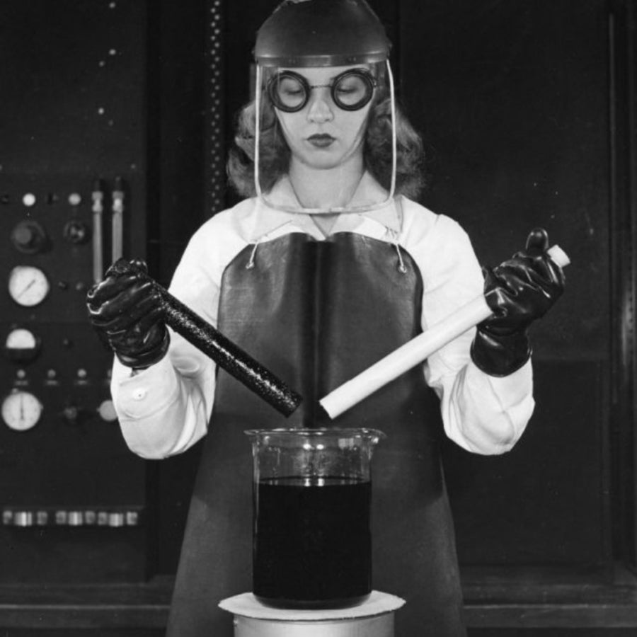 Image vintage en noir et blanc d'une femme en blouse de laboratoire manipulant deux tubes à essai au-dessus d'un bécher, illustrant la fabrication artisanale et ancestrale des hydrolats.